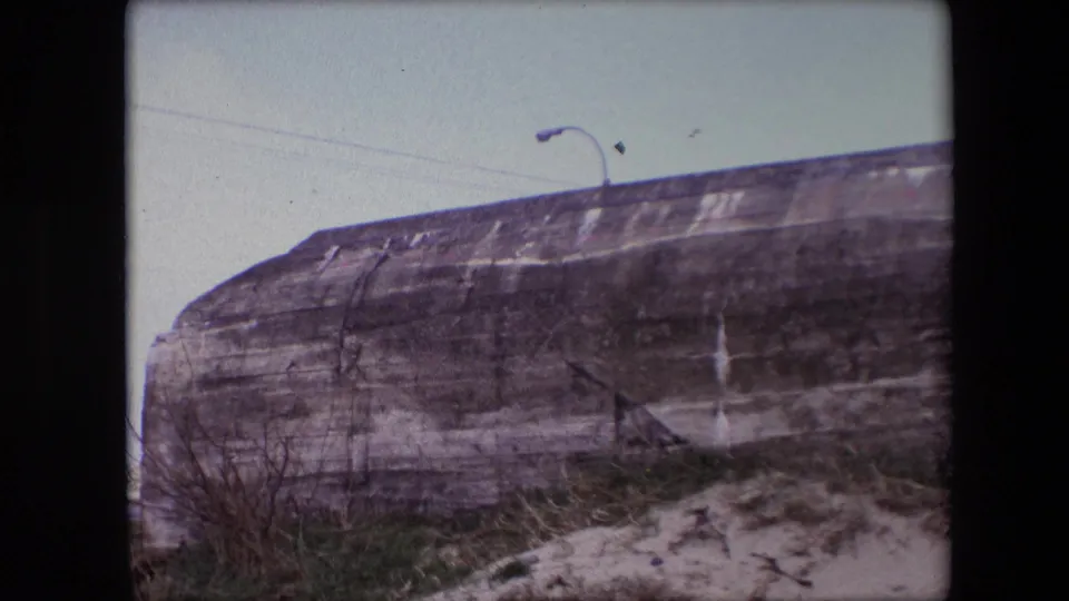 1967 Calais 8mm frame showing a weathered concrete bunker wall, sandy foreground, sparse grass, and a curved streetlamp rising above the top edge.