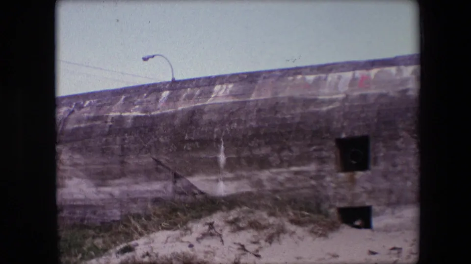 1967 Calais 8mm frame with a closer view of the weathered bunker face, showing two dark square apertures, sandy ground, grass, and streaked concrete.