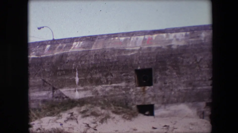 1967 Calais 8mm frame showing the bunker facade with dark square openings, a sandy base, sparse vegetation, and the curved streetlamp visible above.