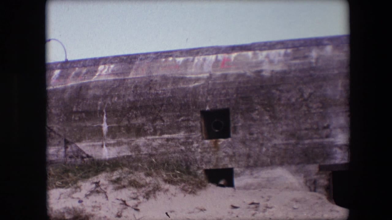 1967 Calais home-movie frame showing a weathered concrete bunker wall partly buried in sand, with square openings, scrub grass, and a pale sky.