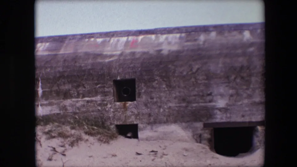 1967 Calais frame of a long weathered concrete bunker, showing small openings, sandy ground, scrub vegetation, and a larger dark entrance on the right.