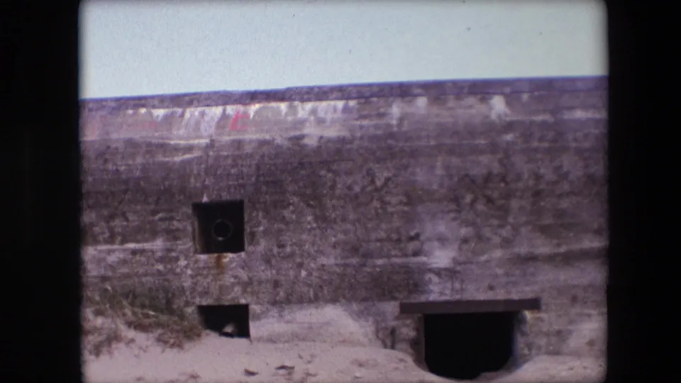 1967 Calais 8mm frame showing the bunker wall closer in view, with a dark square aperture, lower opening, sand, grass, and part of a larger entrance.