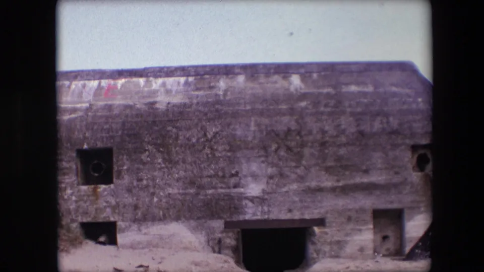1967 Calais frame showing the right side of the weathered bunker, including small openings, a larger dark recess, a doorway-like section, sand, and pale sky.