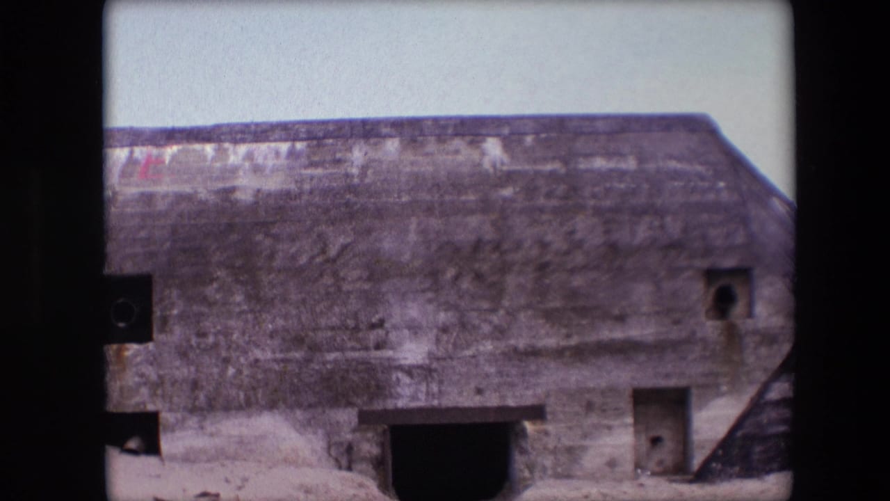 1967 Calais home-movie frame showing a weathered concrete beach bunker, small apertures, a dark entrance, pale sky, and sand against the facade.