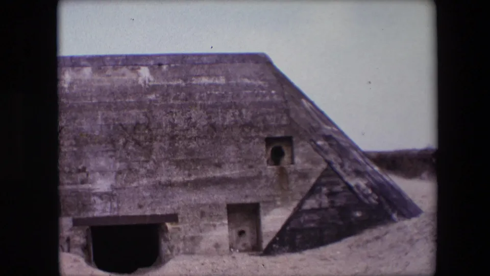 1967 Calais home-movie frame showing the beach bunker from a closer angle, with sand piled along the base and small apertures in the concrete wall.