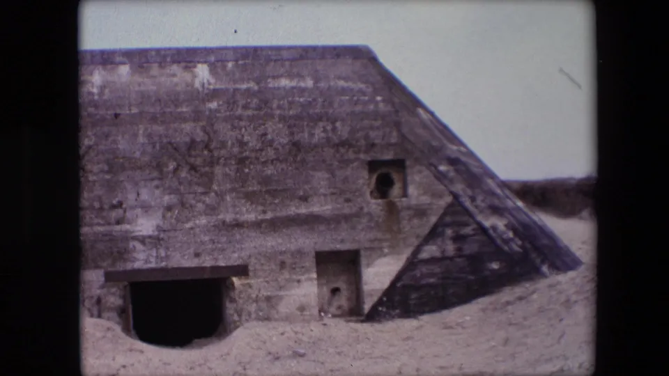 1967 Calais frame showing the bunker centered on the sandy beach, with weathered concrete, a dark entrance, small square openings, and a sloped side wall.