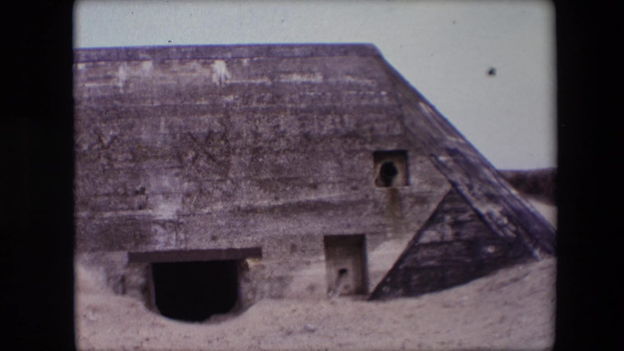 1967 Calais home-movie frame showing a weathered concrete bunker exterior with a dark arched opening, square apertures, sand at the base, and a sloped roof-like section.