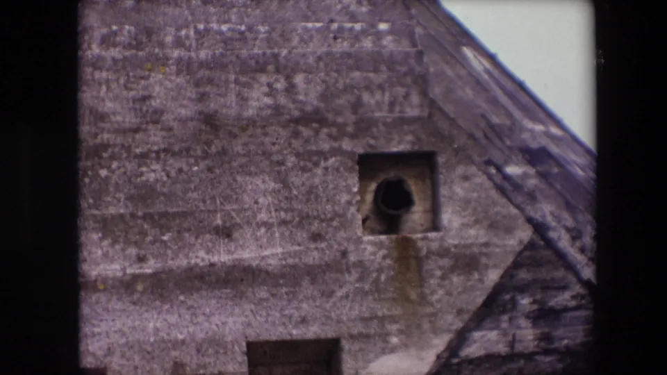 1967 Calais frame centered on a square bunker opening with a dark cylindrical pipe-like form inside, stained concrete beneath it, and part of the sloped structure at right.