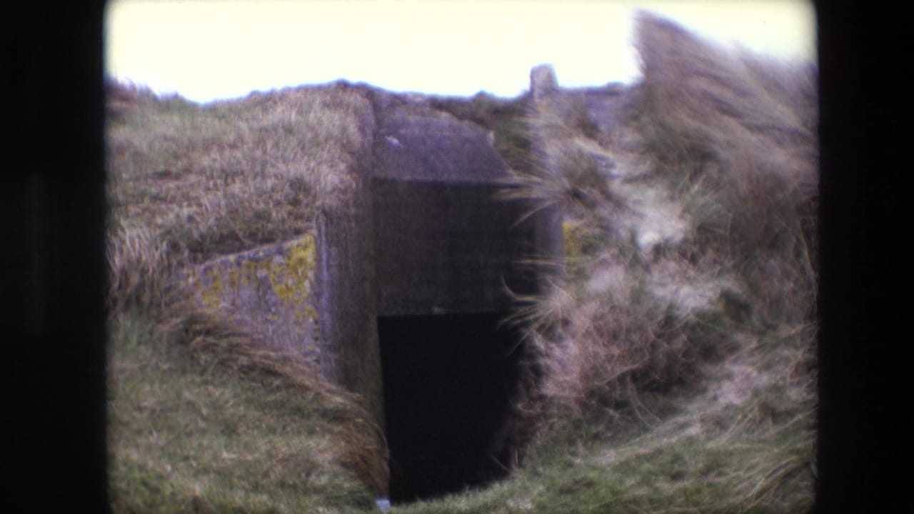 1967 Calais 8mm frame showing a dark concrete bunker-like entrance set into windblown dune grass with lichen on the side wall.