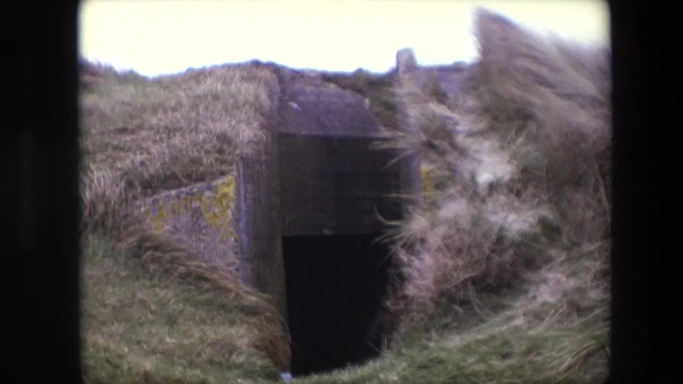 1967 Calais 8mm frame showing a dark concrete bunker-like entrance set into windblown dune grass with lichen on the side wall.