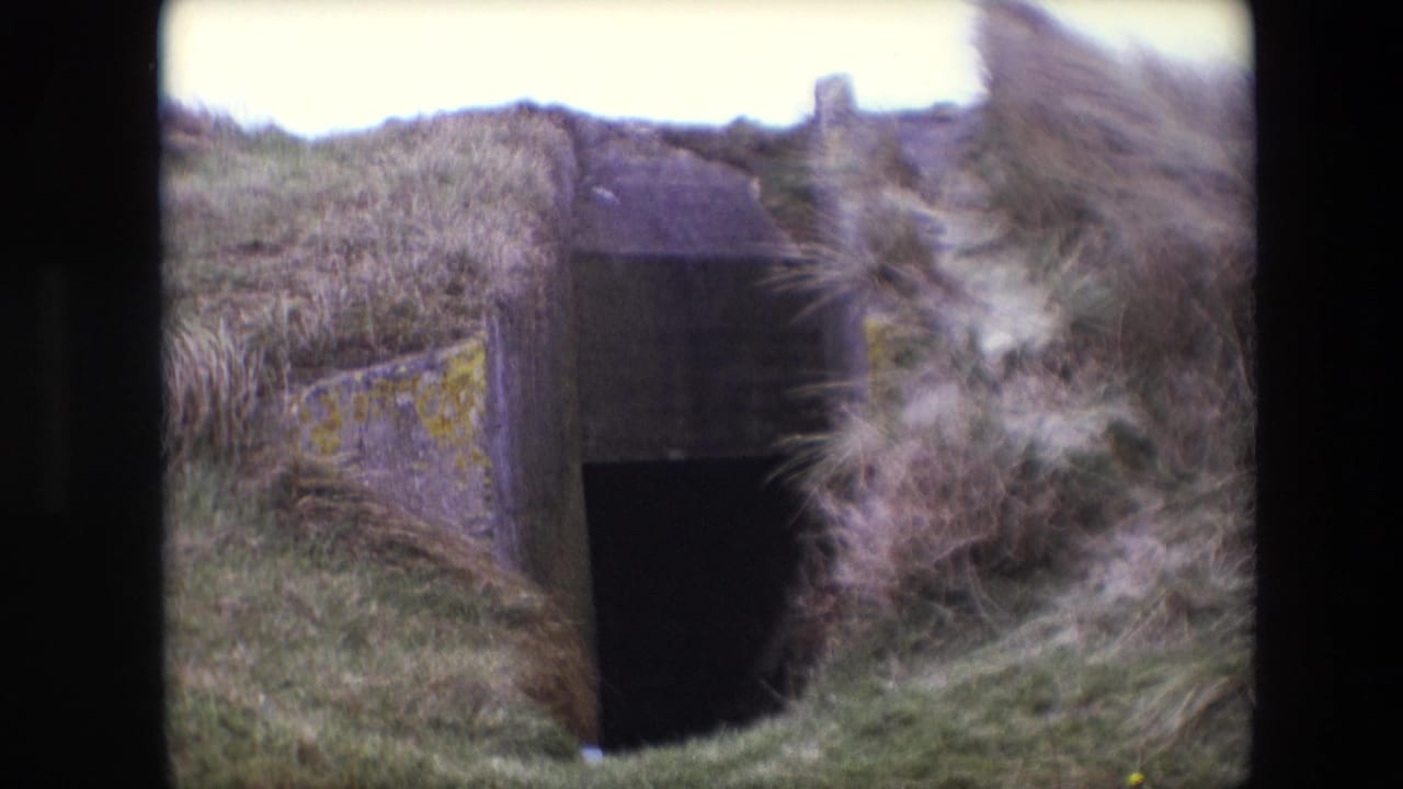 1967 Calais frame with the bunker-like entrance centered between weathered concrete and tall grass bending across the foreground.