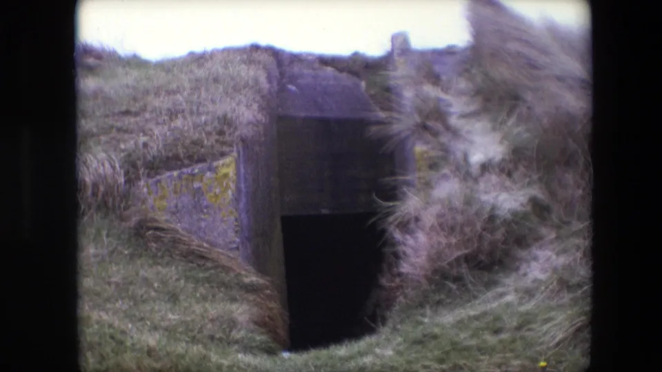 1967 Calais 8mm still showing a low concrete entrance partly obscured by dune grass and soft motion blur from wind or handheld filming.