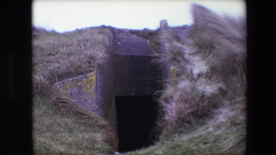 1967 Calais home-movie frame showing a dark rectangular entrance beneath overgrown grass and a stained concrete wall.
