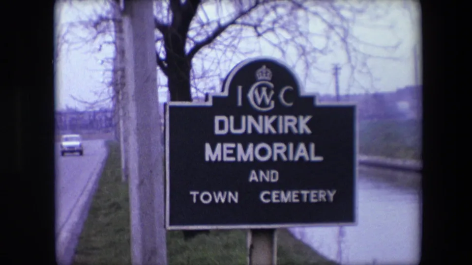 1967 8mm frame from the Calais, France clip showing a roadside sign for Dunkirk Memorial and Town Cemetery near bare trees, a road, and a waterway.