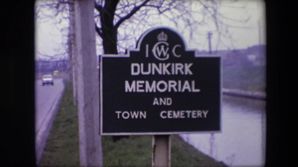 Closer 1967 8mm frame from the Calais, France clip showing the Dunkirk Memorial and Town Cemetery sign beside a road, trees, and a canal or river edge.