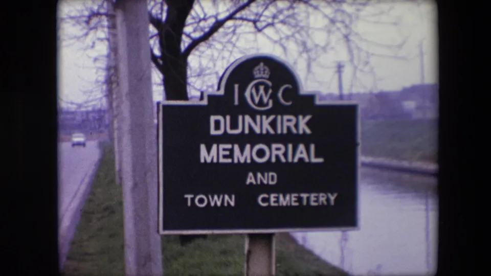 1967 8mm frame showing the Dunkirk Memorial and Town Cemetery sign beside a quiet road and canal, with a small car visible in the distance.