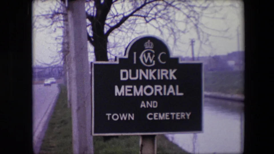 1967 8mm frame with a close view of the black Dunkirk Memorial and Town Cemetery sign, roadside pole, canal edge, and bare trees.