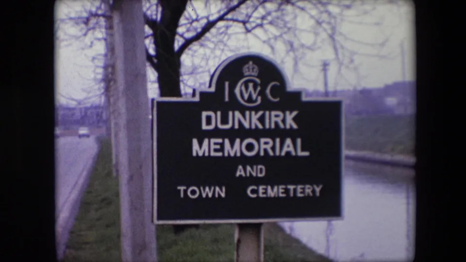 1967 8mm frame showing the Dunkirk Memorial and Town Cemetery sign at the roadside, with canal water and winter-bare trees in the background.