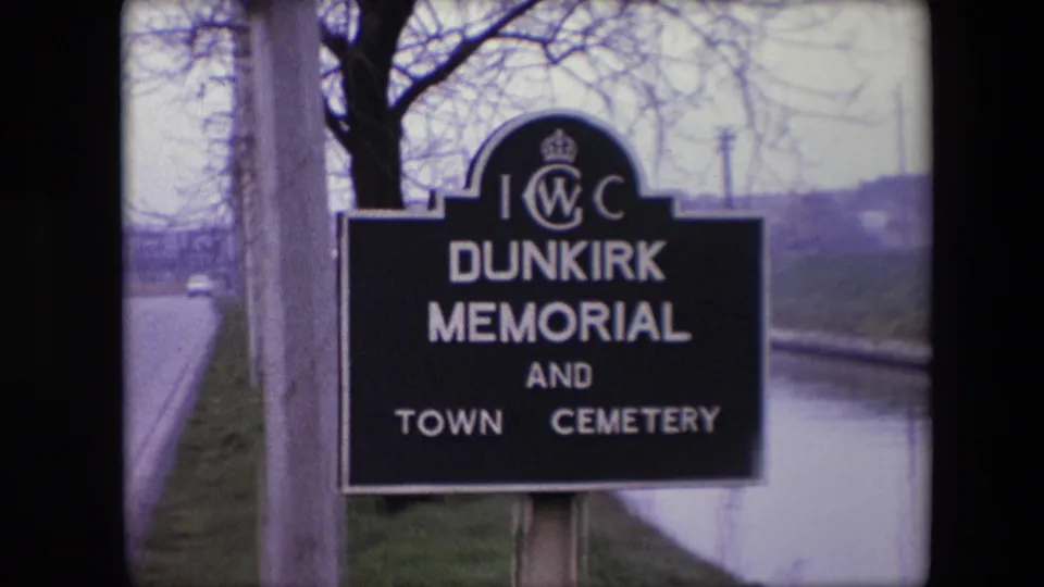 1967 8mm frame from Calais, France showing the Dunkirk Memorial and Town Cemetery sign beside a quiet roadside with bare trees in the background.