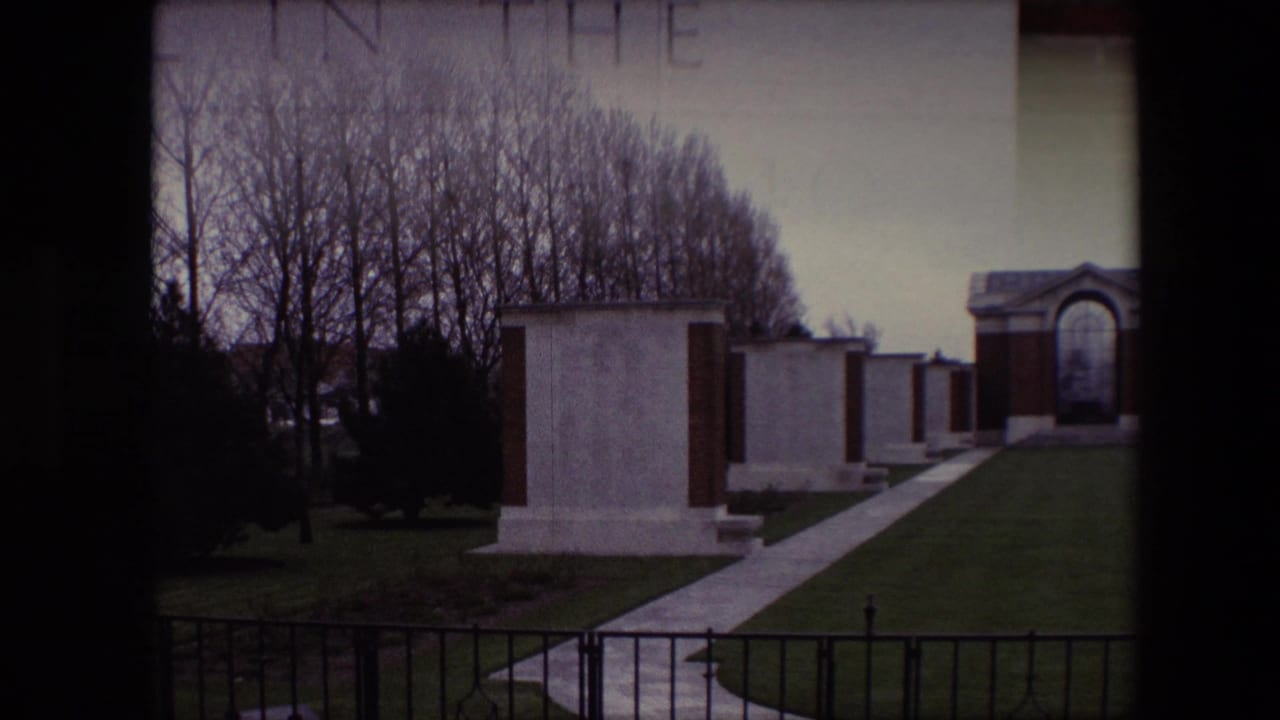 1967 Calais, France 8mm frame showing white gravestones beside a paved pathway, green lawn, bare trees, a black fence, and an arched memorial structure in the distance.