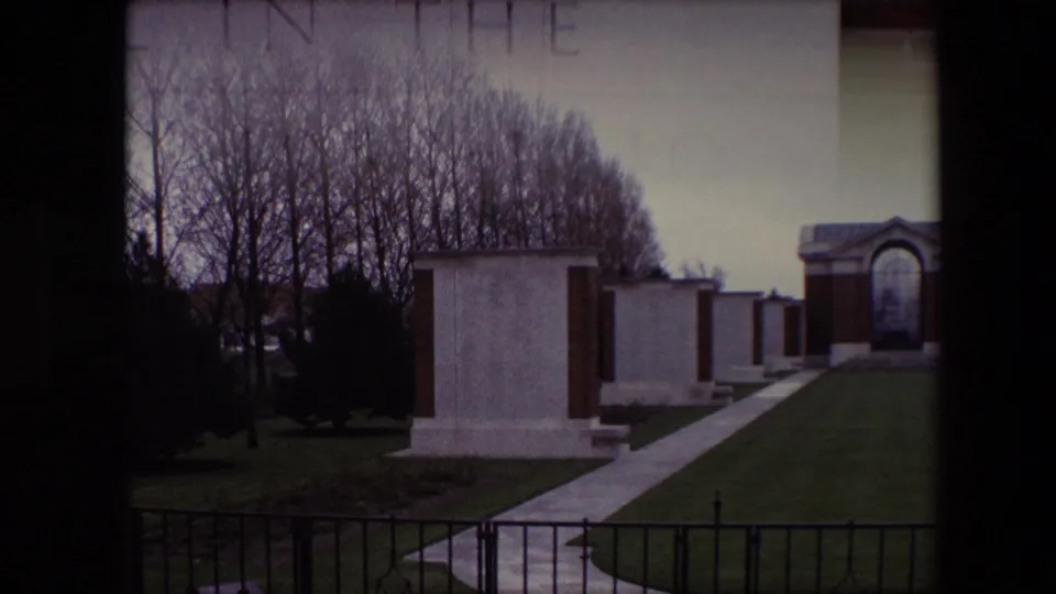 1967 Calais, France 8mm frame showing white gravestones beside a paved pathway, green lawn, bare trees, a black fence, and an arched memorial structure in the distance.