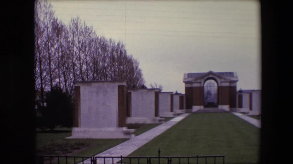 1967 Calais, France 8mm frame showing a closer visitor view of white grave markers, a paved walkway, clipped grass, bare trees, and the arched memorial structure at the end of the lawn.