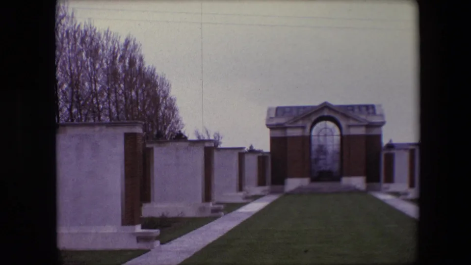 1967 Calais, France 8mm frame centered more tightly on an arched memorial structure, with a sloping lawn, paved paths, pale gravestones, bare trees, and a foreground fence.