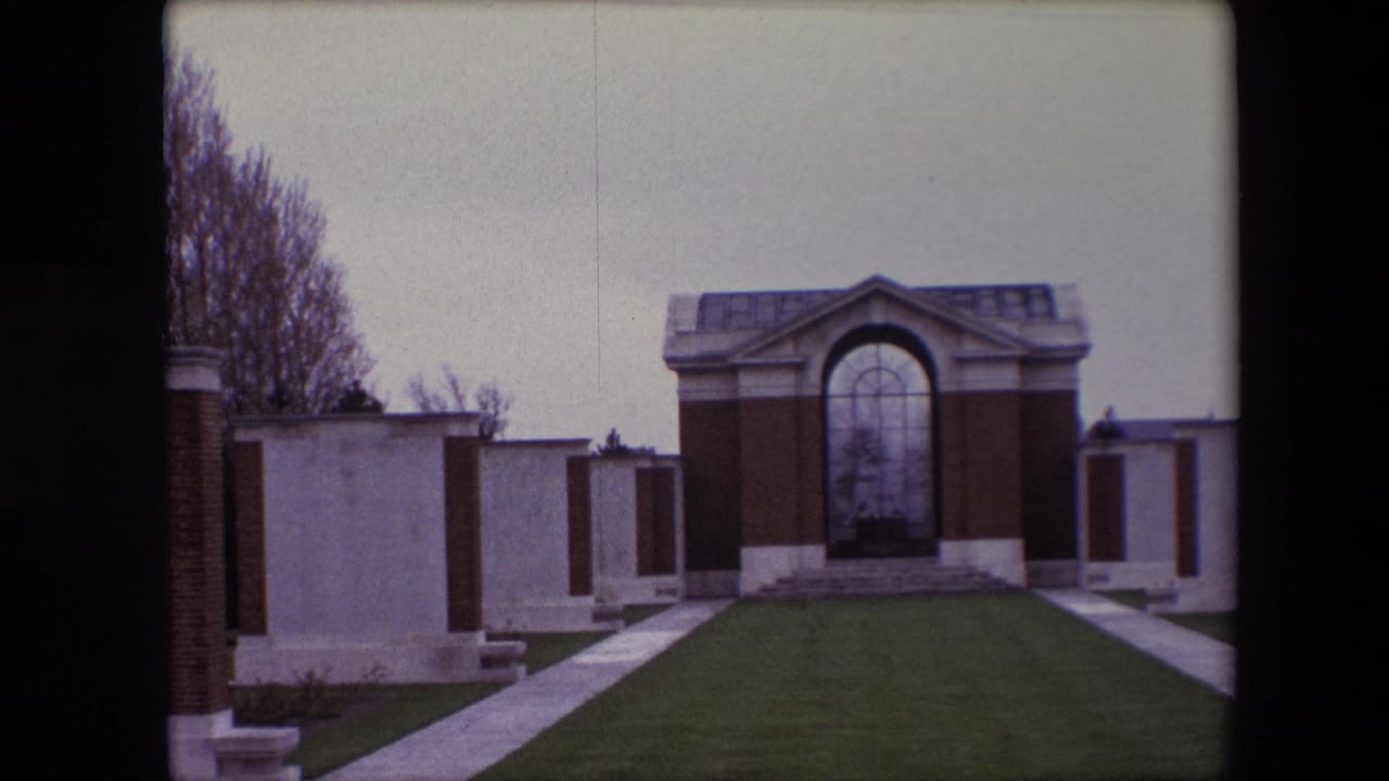 Wide 1967 Calais cemetery view showing a brick-and-stone memorial structure with an arched glass panel, lawn, walkway, and white headstones at the sides.