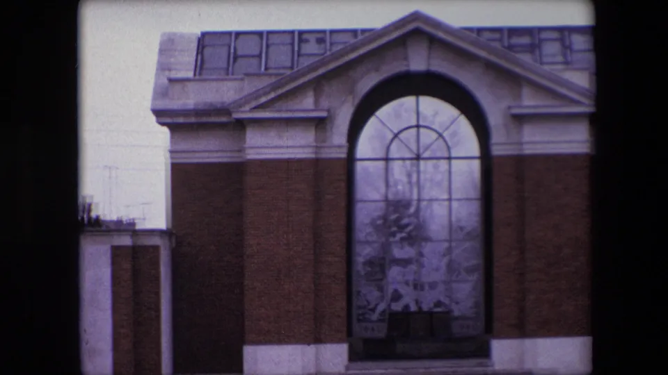 Tight 8mm frame of the Calais memorial facade, showing brickwork, pale stone trim, and the large arched glass panel with decorative figures or scene detail visible through the soft focus.