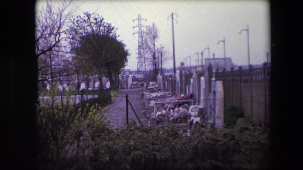 1967 Calais home-movie frame showing a quiet cemetery path bordered by gravestones with flowers, bare trees, lamp posts, and utility pylons in the distance.