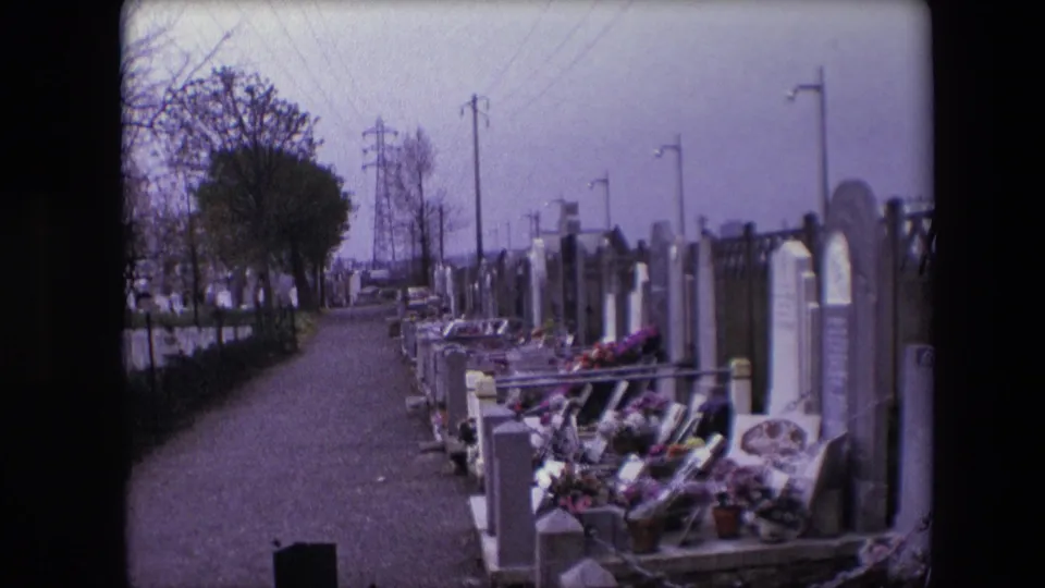 1967 Calais 8mm frame showing a cemetery path with flowered graves and upright markers on the right, trees and utility pylons receding into the background.