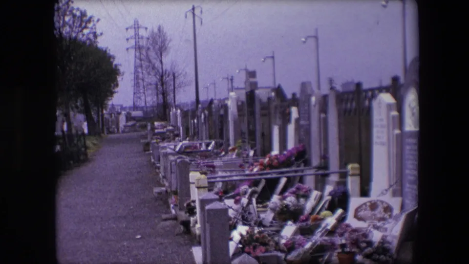 1967 Calais home-movie frame with flowered graves and headstones along a cemetery path, framed by bare trees, lamp posts, and distant utility pylons.