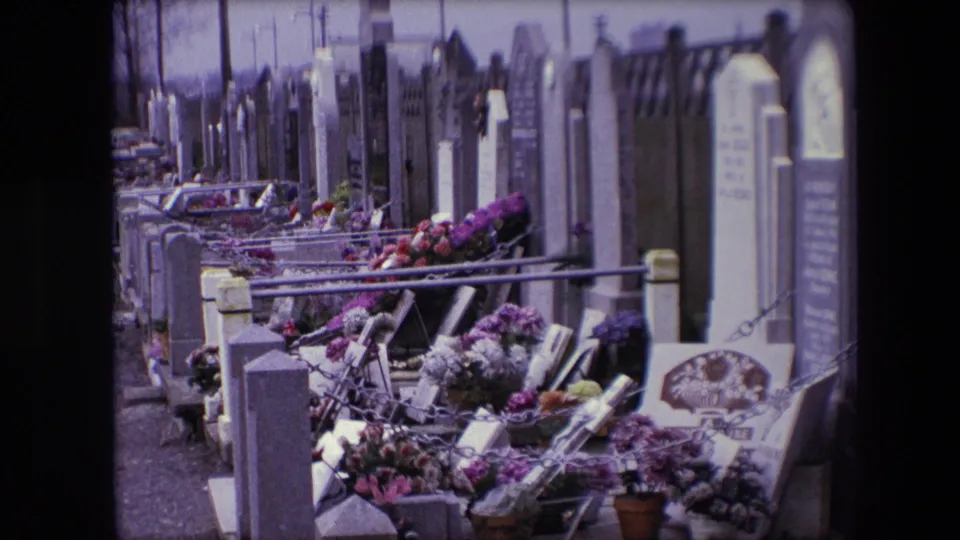 Flower-covered grave plots and upright headstones in a Calais cemetery, filmed from a visitor's moving perspective in 1967.