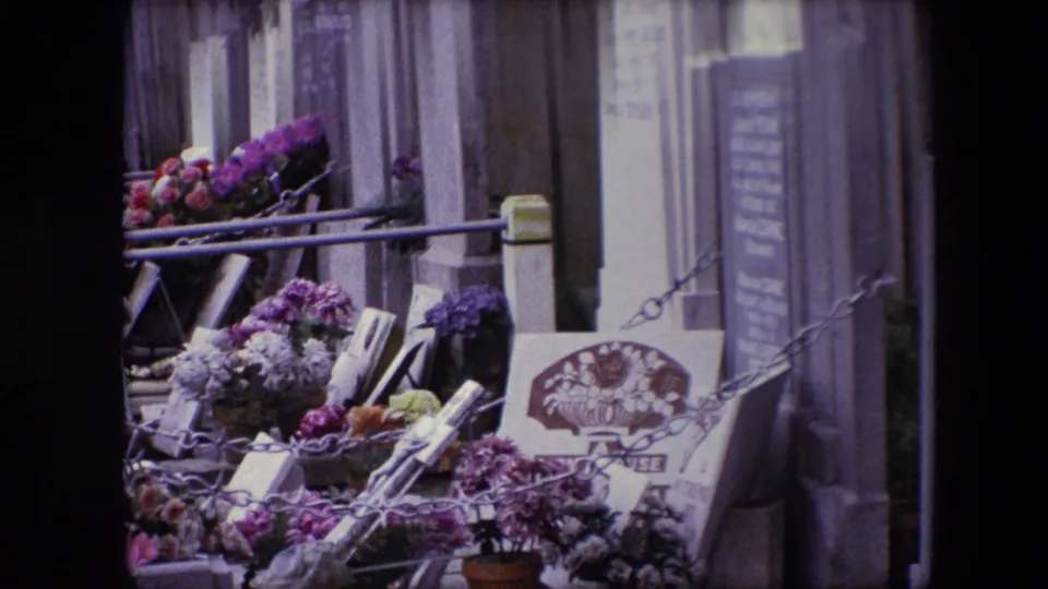 End frame from the 1967 Calais cemetery footage, showing floral tributes, metal railings, and partially legible grave markers in soft 8mm focus.