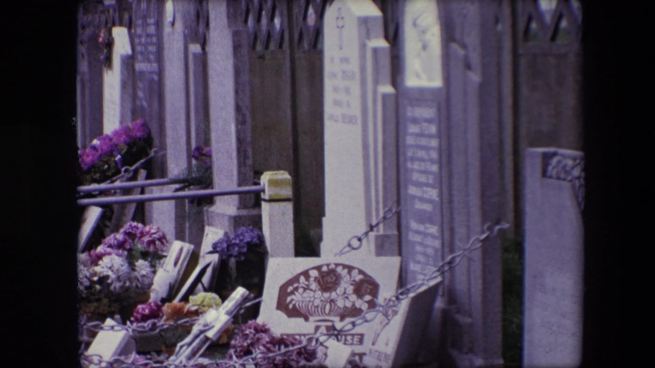 1967 Calais 8mm frame showing cemetery plots with purple and white flowers, upright grave markers, metal railings, chains, and a wall in the background.
