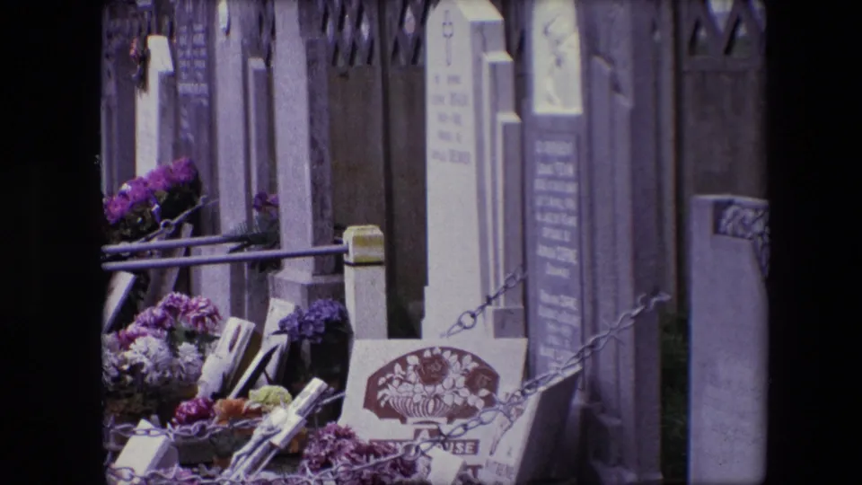 1967 Calais 8mm frame showing cemetery plots with purple and white flowers, upright grave markers, metal railings, chains, and a wall in the background.