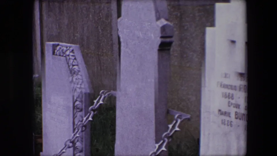 1967 Calais 8mm frame with a close view of grave markers, a chain railing, grass, a decorative stone edge, and a partly readable inscription.