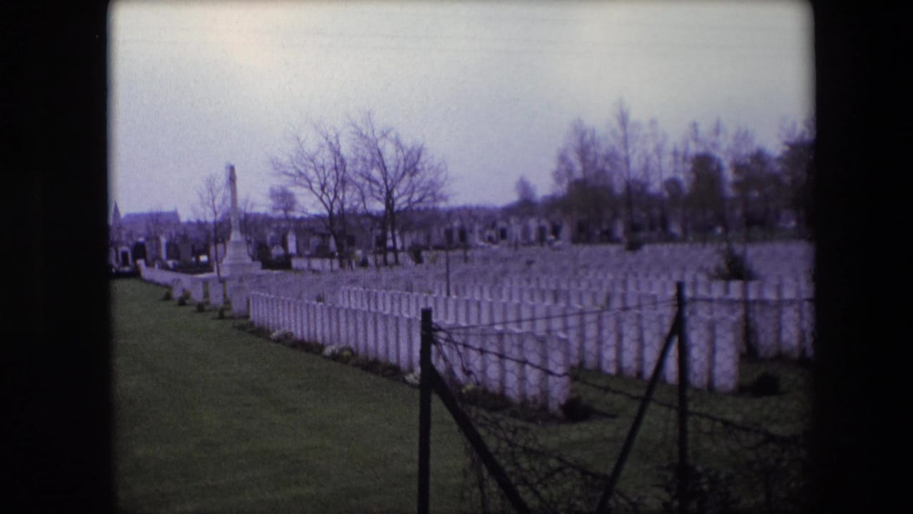 Calais, France, 1967 8mm frame showing rows of white cemetery headstones, a dark foreground fence, bare trees, and a tall stone monument under an overcast sky.