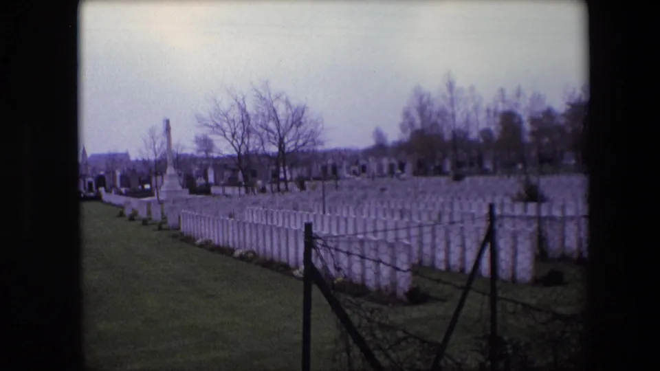 Calais, France, 1967 8mm frame showing rows of white cemetery headstones, a dark foreground fence, bare trees, and a tall stone monument under an overcast sky.