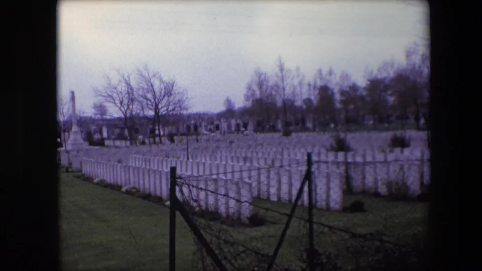 Calais, France, 1967 8mm frame looking across uniform white headstones from behind a dark wire fence, with leafless trees and distant grave markers in the background.