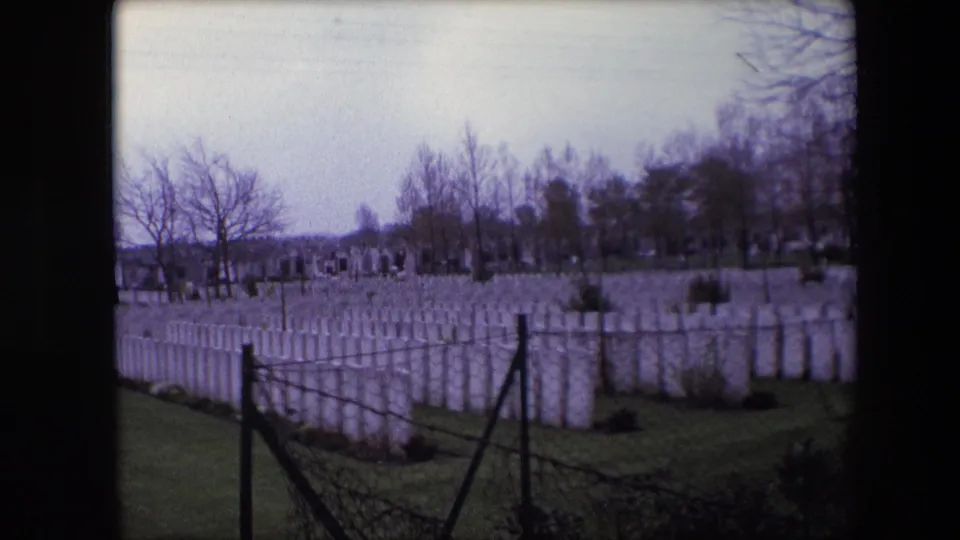 Calais, France, 1967 8mm frame with the camera angled across a maintained cemetery lawn, showing many white headstones, a dark fence, and bare trees beyond.