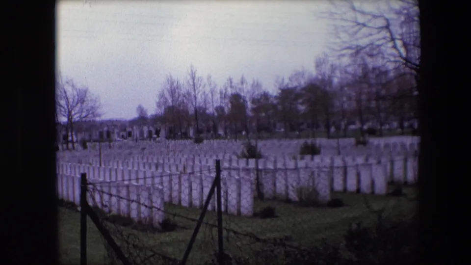 Calais, France, 1967 8mm frame showing a wide view of white headstones across a green cemetery lawn, framed by dark fence posts and winter trees.