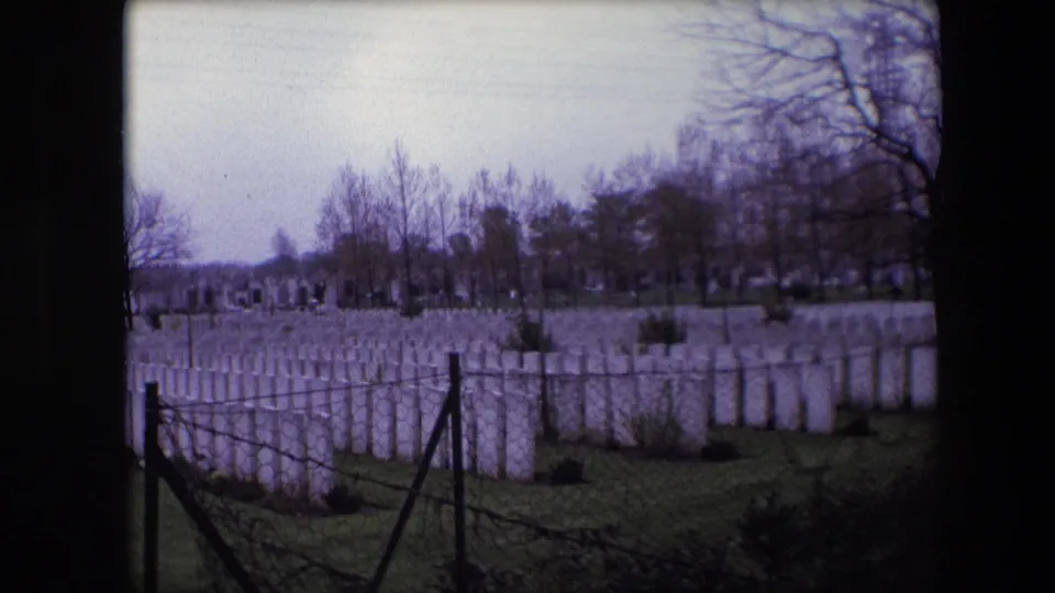 1967 Calais, France 8mm frame showing rows of white military cemetery headstones behind chain-link fencing and barbed wire under bare trees.