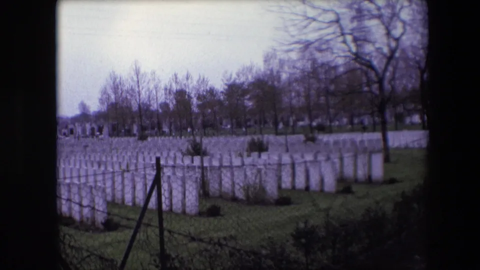 1967 Calais, France 8mm frame with uniform white headstones across a grassy military cemetery, seen past chain-link fencing and bare trees.