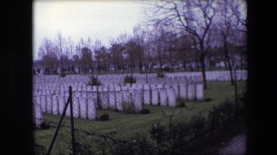 1967 Calais, France 8mm frame showing a panned view of white military cemetery headstones, a chain-link fence, barbed wire, and leafless trees.