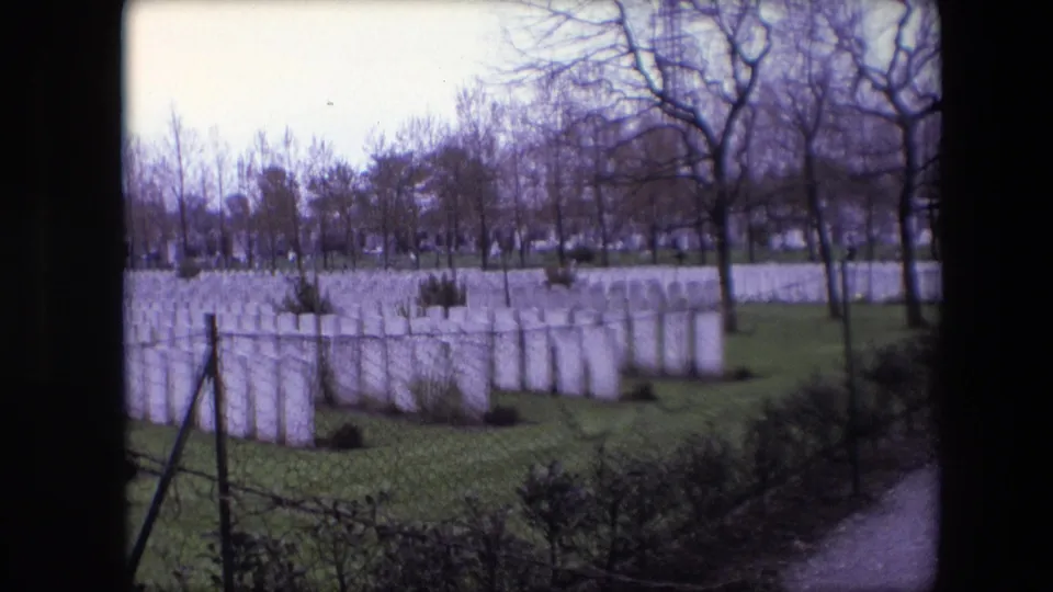 1967 Calais, France 8mm frame showing rows of white cemetery headstones behind wire fencing, with shrubs, bare trees, and a path along the right edge.