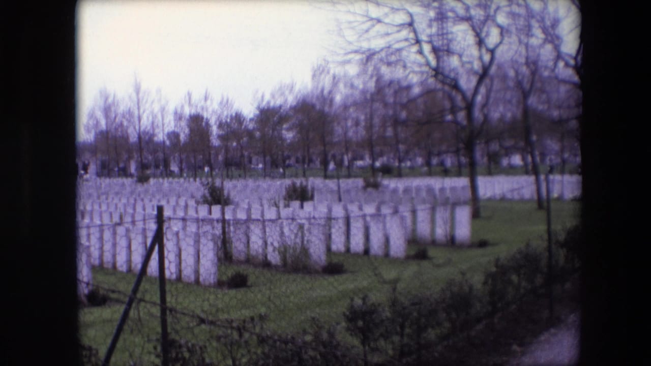 1967 Calais home-movie frame showing rows of white military cemetery headstones beyond a chain-link fence, with grass and bare trees under a pale sky.