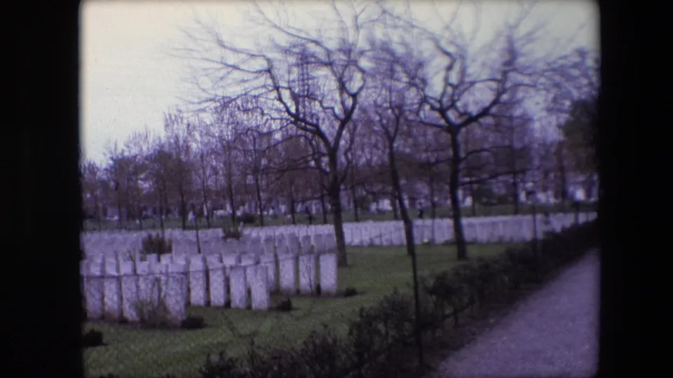 1967 Calais 8mm still showing white military cemetery headstones beside a paved path, with chain-link fencing, shrubs, grass, and leafless trees.