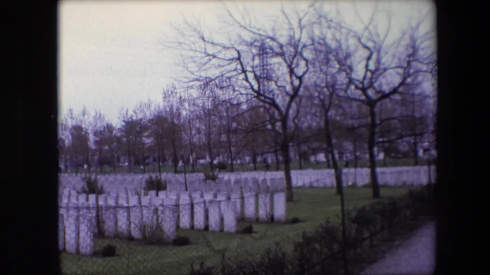 1967 Calais, France 8mm frame showing cemetery rows, leafless trees, a clipped lawn, low hedging, and a pale path at the right.