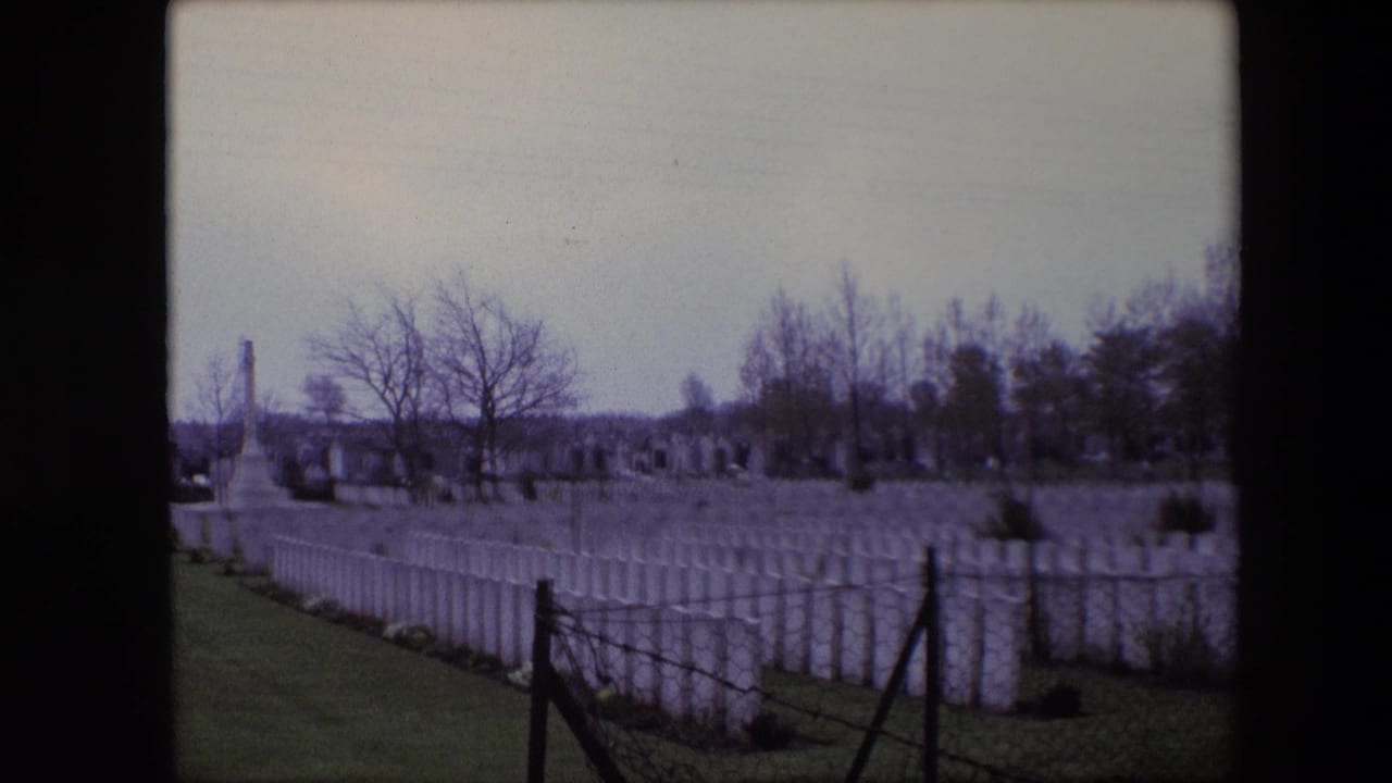 1967 Calais 8mm frame showing rows of white military cemetery gravestones, a grassy path, bare trees, and a distant stone memorial under gray sky.
